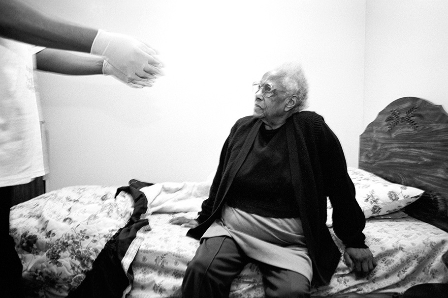 An elderly woman sits on her bed, while her caregiver reaches out to her. 