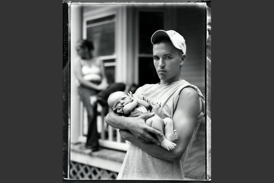 Teenage boy holding a baby with a woman in the background.