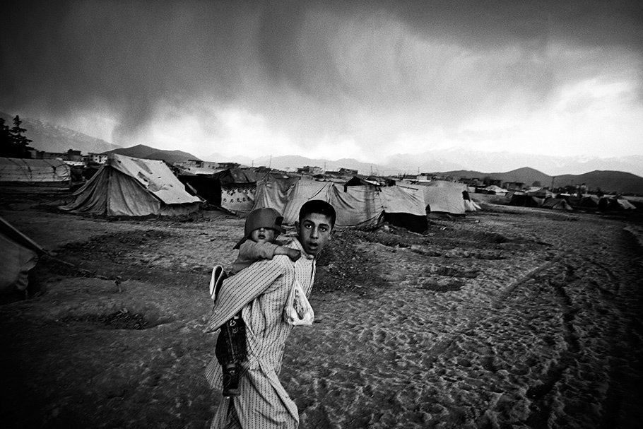Man with child on back, tents in background.