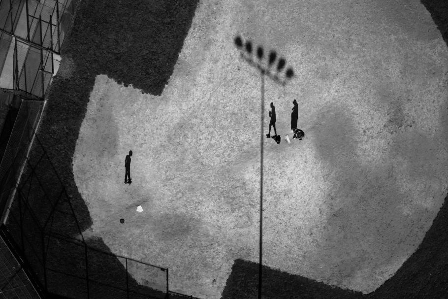 Aerial photograph of three figures and stadium lights casting shadows across a baseball field. 