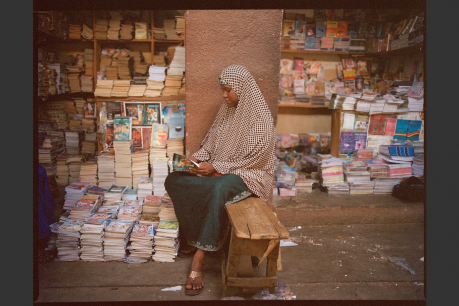 A Nigerian romance novelist reads a book outside a bookstall